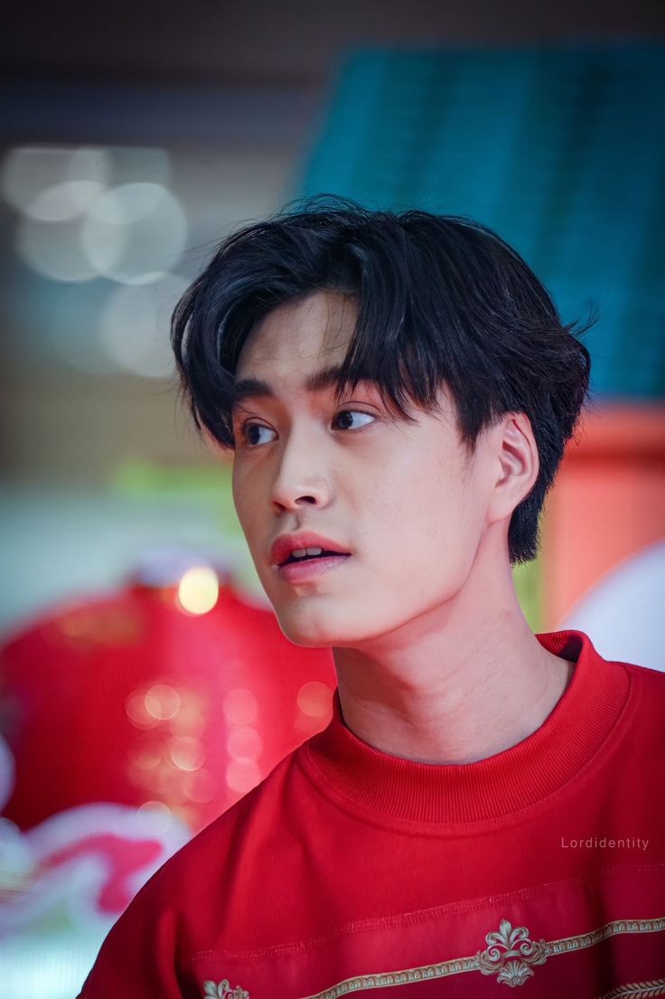 a young asian man wearing a red sweatshirt with gold trim. he has short, dark hair and is looking to the left of the camera with his mouth slightly open as if he's about to speak. the background is blurry and features red decorations, suggesting a festive occasion. the lighting is bright, highlighting the man's features.