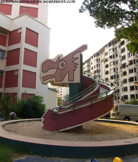a spiral slide that looks like a dragon. the slide is made of red and brown metal with a metal railing. the slide is located on a playground in front of a residential building in singapore.  the slide is surrounded by sand and a circular patio area.  the surrounding buildings are in the background.