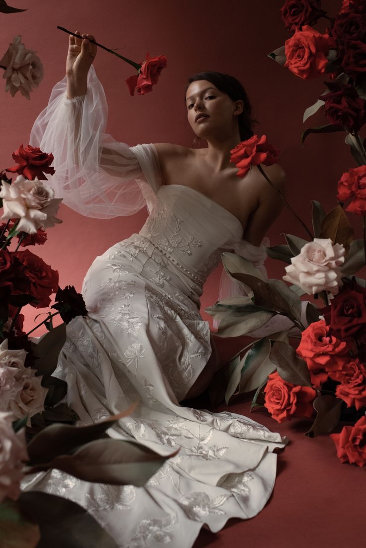 a beautiful bride in a white dress stands in a studio, surrounded by red roses. she is looking at the camera with a gentle expression. the dress is a fitted bodice with a long flowing skirt and features lace.  the flowers are arranged in a way that creates a romantic and dreamy atmosphere.  she is holding a red rose in her right hand.  the  lighting is soft and natural, creating a warm and inviting feeling. the background is a red wall, which contrasts nicely with the white of the dress. the overall effect of the image is one of elegance and beauty.