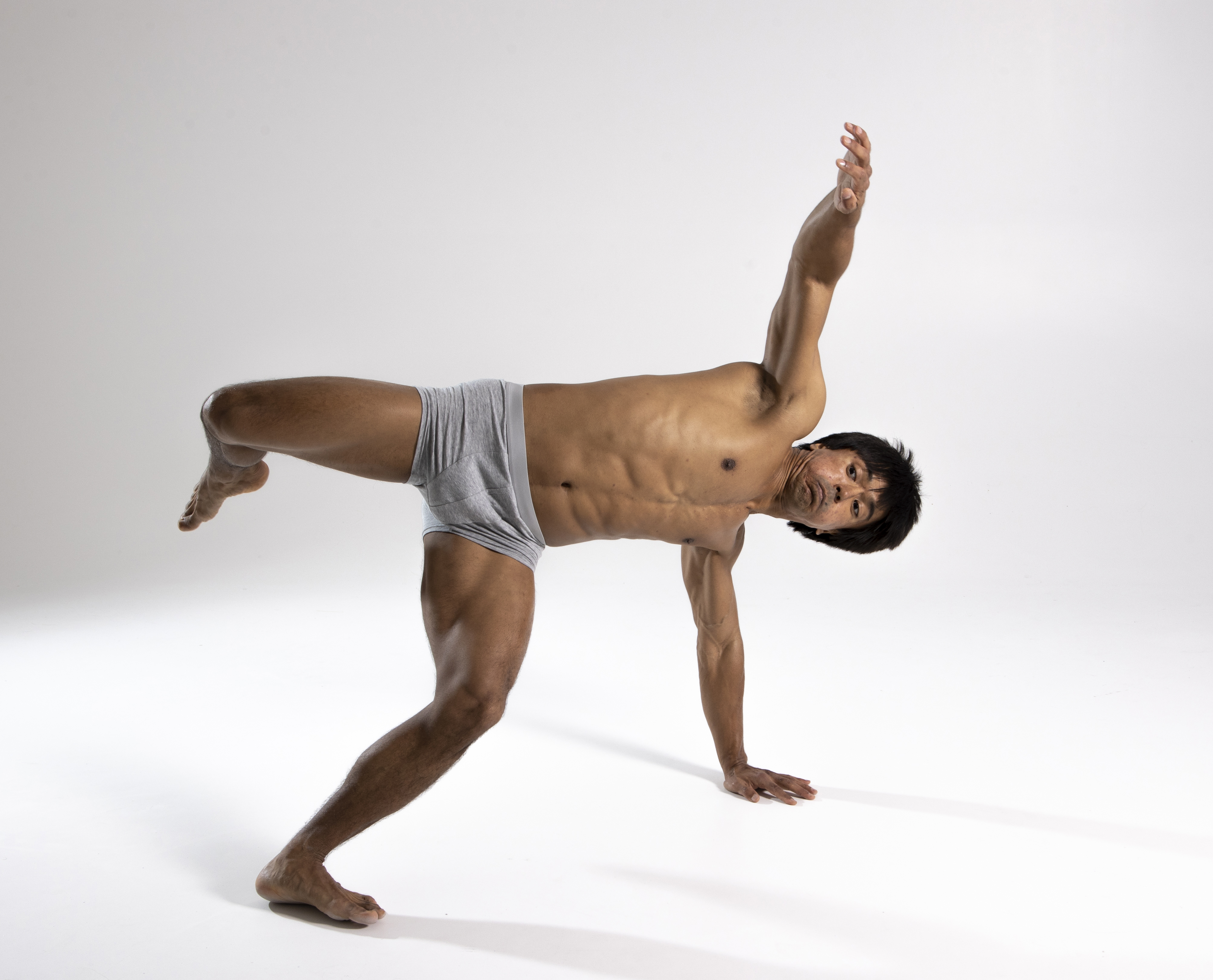 a shirtless man in gray briefs is posed in a studio shot. he is in an athletic pose, standing on one hand, with one leg lifted and one arm raised. he appears to be a dancer or fitness enthusiast, showcasing his muscular physique and balance. he is bare feet.