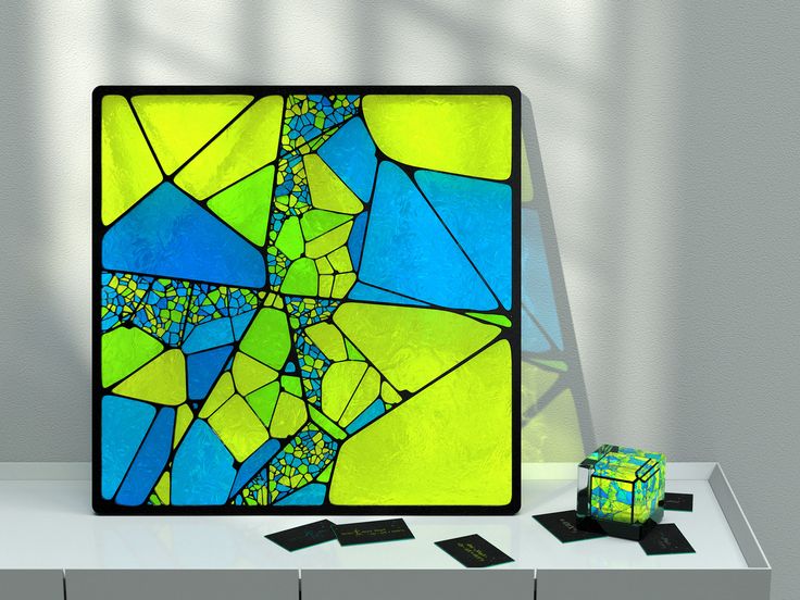 the image shows a square abstract art piece with a black frame leaning against a wall on a white shelf. the art consists of various geometric shapes in blue, green, and yellow. beside the artwork is a matching cube surrounded by black business cards. a shadow is cast on the wall behind the artwork.