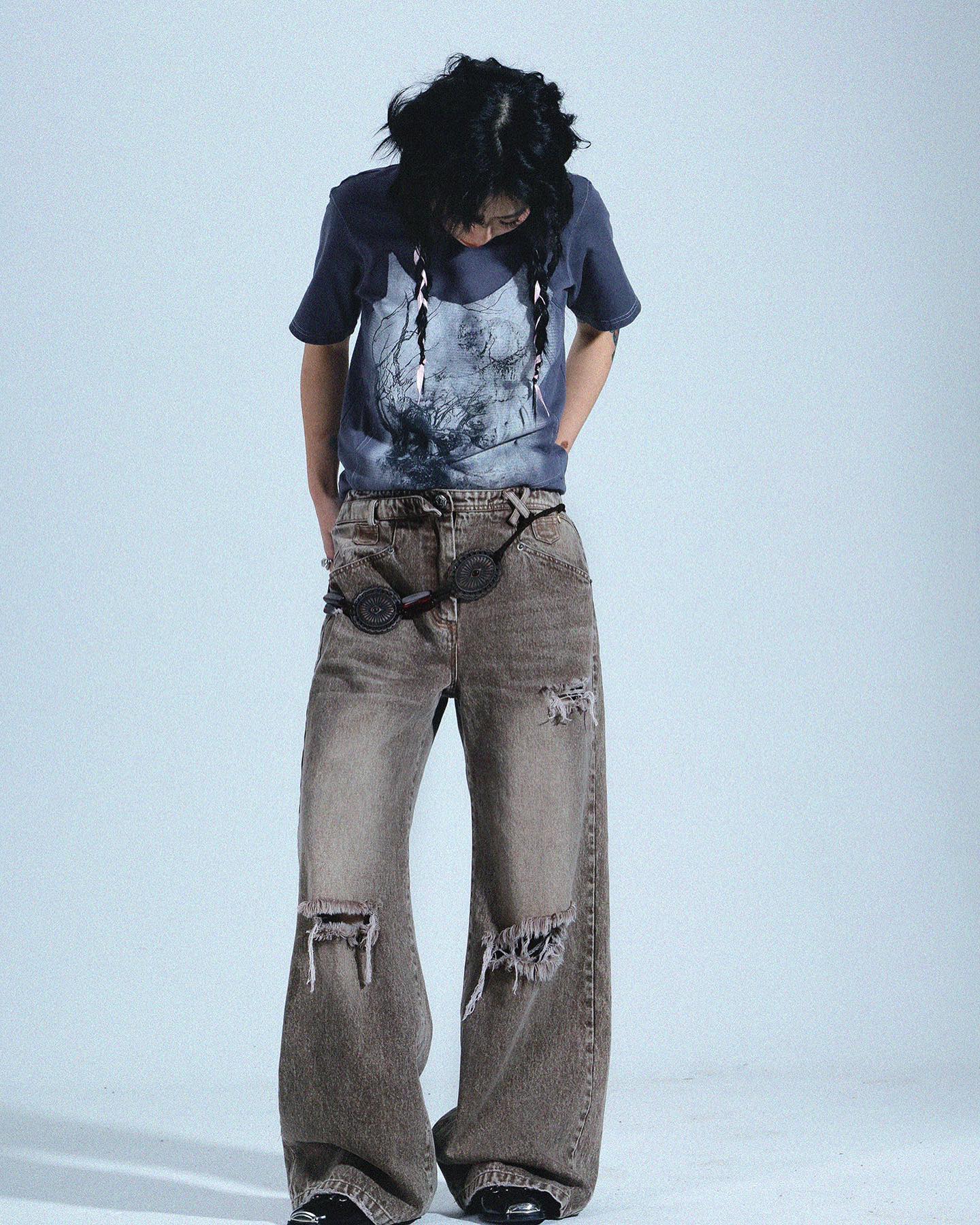 a fullbody shot of a young woman posing in a studio setting against a light gray backdrop.  she is wearing a dark gray tshirt featuring a graphic print, a pair of wideleg, distressed, grayishbrown jeans, and a darkcolored belt with circular accents. her dark hair is styled in long braids that hang down her back. the overall style is casual and slightly grunge or alternative.  the focus is on the clothing and the model's pose.