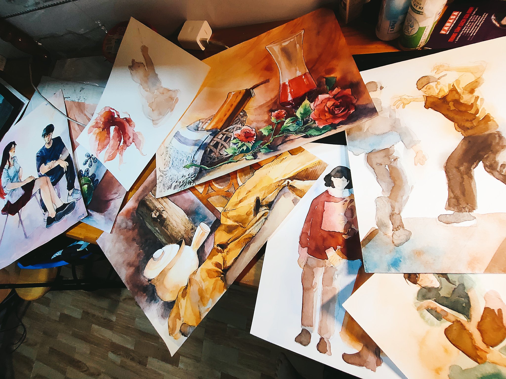 highangle, closeup shot captures a collection of watercolor paintings scattered across a wooden table. the paintings showcase a diverse range of subjects, from still life arrangements featuring vases and flowers to human figures in various poses and settings. the artist's workspace is evident, with paint splatters and the slightly disheveled arrangement of the artwork suggesting a creative and dynamic process.  the setting appears to be a home or studio, with a glimpse of a floor and other household elements visible in the background. the overall impression is one of artistic energy and productivity.