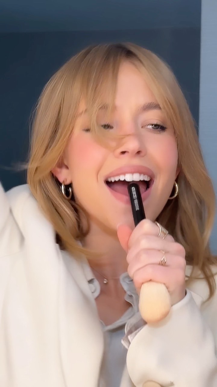 loseup view of a young woman with blonde hair and bangs. she is smiling and holding a makeup brush, appearing to be applying makeup. she's wearing a white jacket and has gold earrings and rings. the overall mood is bright and cheerful.