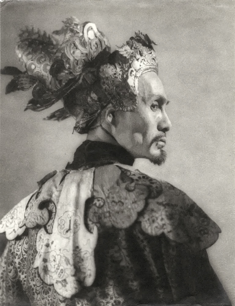 the image is a black and white, vintagestyle photograph of a man, likely an actor, dressed in an elaborate chinese opera costume.  he's wearing a large, ornate headdress that extends over his shoulders, filled with intricate details. the costume itself is richly decorated with embroidery and details on the shoulders and cape area. his facial expression is serious, possibly pensive. the photograph is taken from a slightly behind threequarter profile, highlighting the details of his clothing and headdress. the background is plain and unfocused, emphasizing the subject. the overall style suggests a historical or theatrical setting.