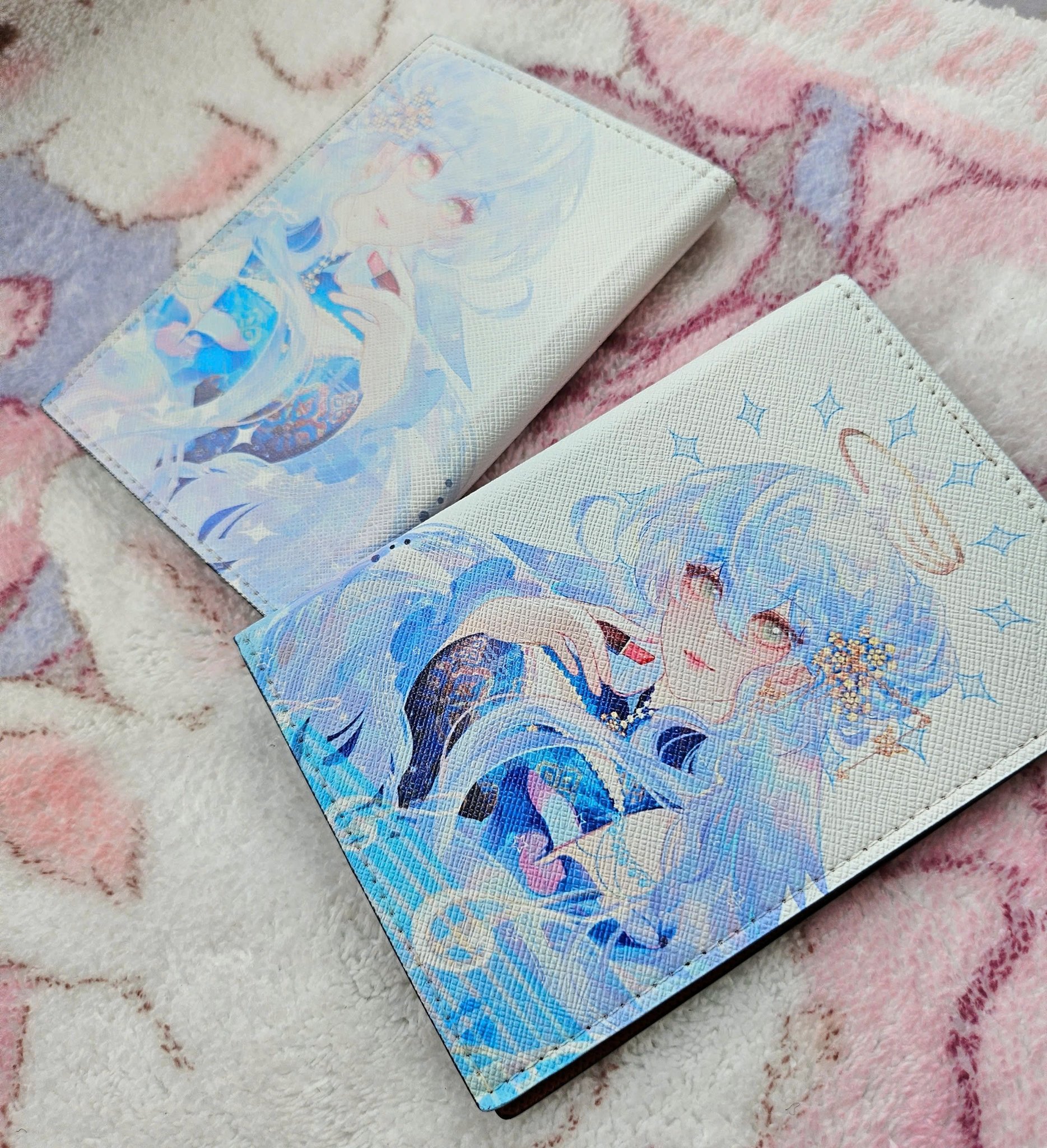 the image shows two white wallets lying on a soft pink and white textured blanket. both wallets feature a different illustration of an animestyle girl with long blue hair. the illustrations are soft, pastel, and feature a dreamy, ethereal quality.  the wallets appear to be made of a smooth, possibly leatherlike material. the design is cute and kawaii.