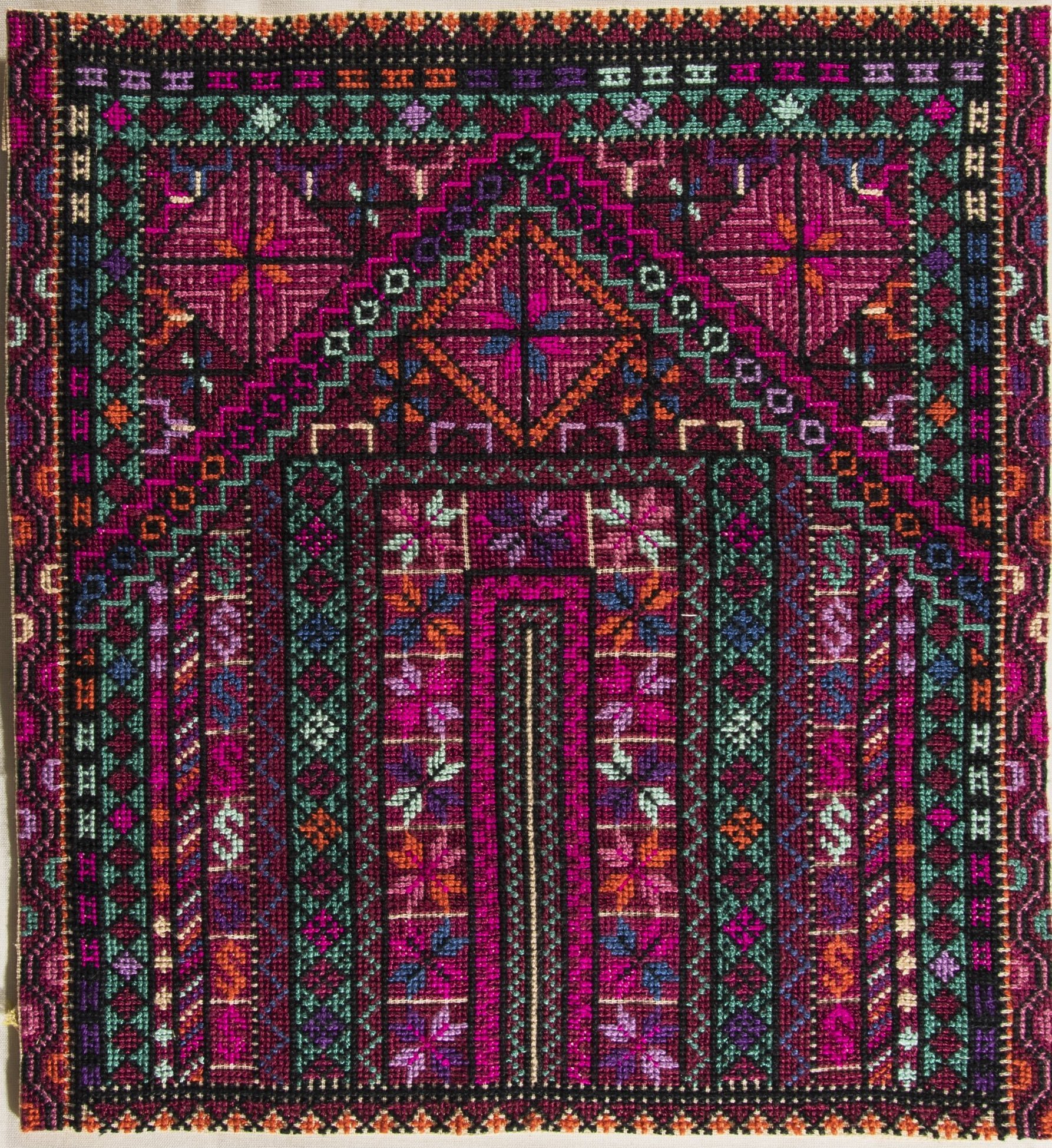 a closeup image of a handstitched textile featuring a complex geometric pattern in shades of pink, purple, orange, green, and black. the design includes repeating motifs and intricate crossstitching, characteristic of traditional middle eastern or palestinian folk art embroidery. the textile has a decorative, handcrafted appearance and resembles a small carpet or rug.