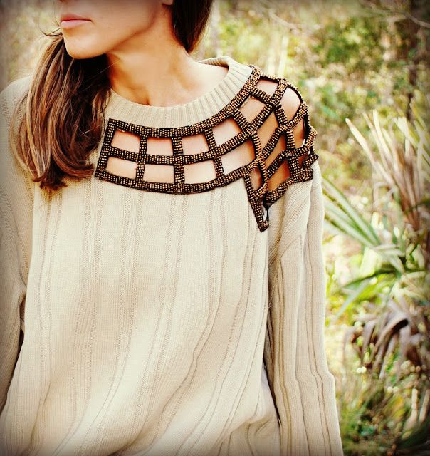 a woman wearing a beige sweater with a unique cutout collar poses for a photograph. the collar is a gridlike design of brown and beige material, exposing sections of skin. the longsleeved sweater features a vertical texture pattern. the background shows blurred green foliage, hinting at an outdoor setting. the woman's face is partially visible, revealing a portion of her hair and her jawline.