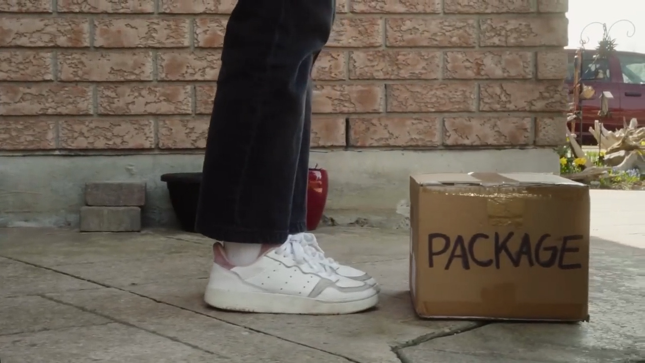 a person wearing black pants and white sneakers stands on a cement sidewalk in front of a brick house. a cardboard box labeled “package” sits on the ground in front of them.