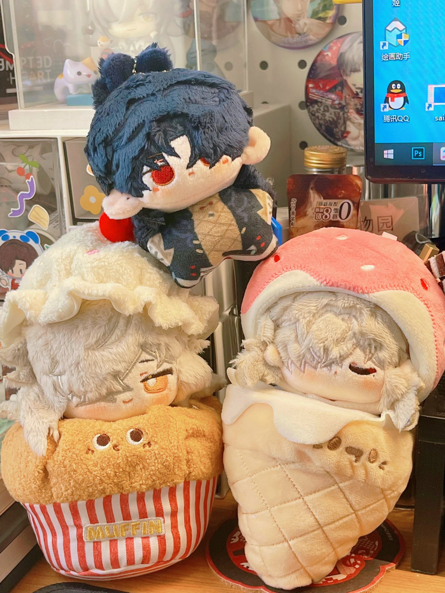 a collection of three plush toys, each resembling an anime character. the one on the left is a small white and brown character dressed in a muffin costume. the middle character is a darkhaired character in a red and blue outfit with a yellow and red flower design. the character on the right is a white and brown character wearing a cone ice cream costume. all three are sitting on a table with a variety of other items in the background, including books and other plush toys.