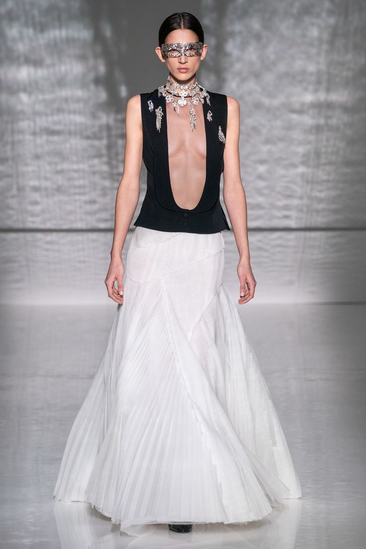 a woman is walking on a runway wearing a long white pleated dress with a sleeveless black top, a silver mask, and an elaborate necklace. the background is a metallic silver.