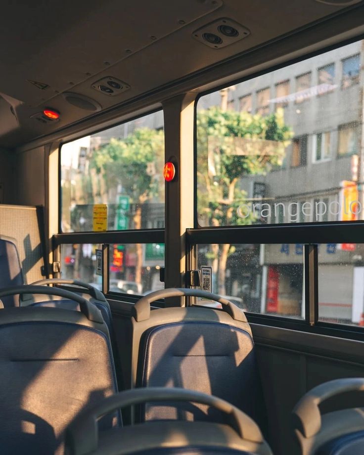 an empty city bus with the windows open and the sunlight streaming through the windows. the bus seats are grey and empty. there are rows of seats in the bus. the bus is moving, and the city is passing by. the shadows of the trees are cast onto the seats.