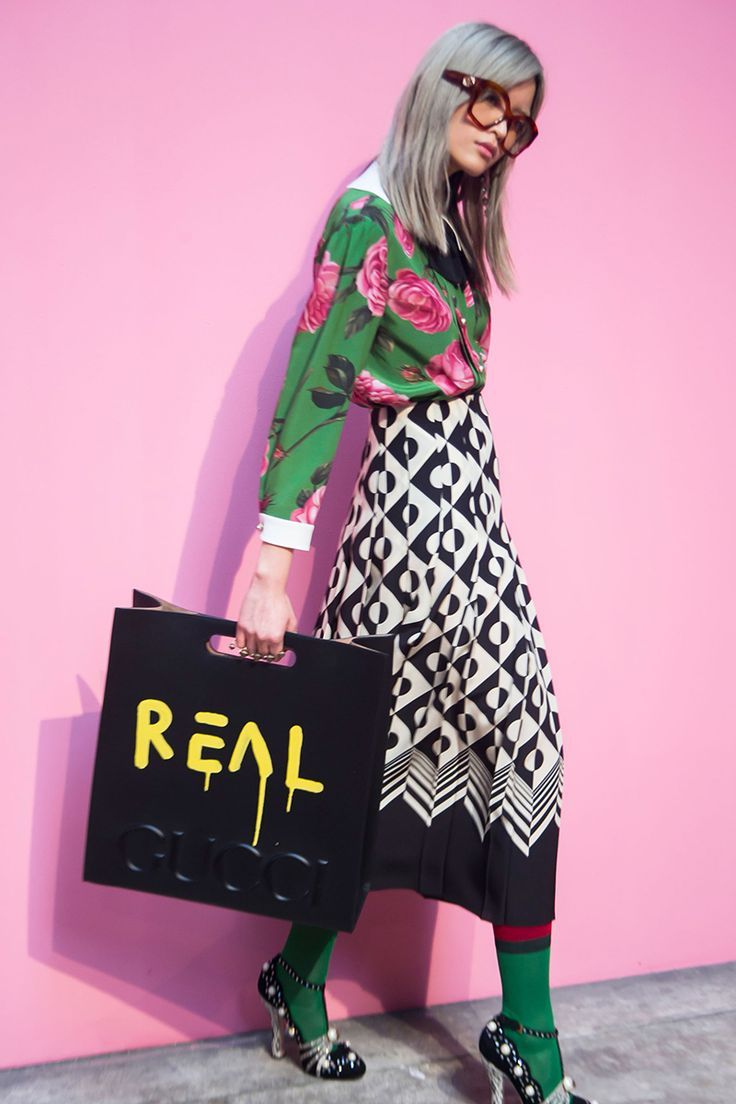 the image shows a fashion model posing against a bright pink background. she's wearing a green floral gucci shirt, a black and white geometric pleated skirt, green socks, and highheeled shoes adorned with pearls.  she carries a black gucci shopping bag with "real gucci" written on it in yellow graffitistyle lettering. the model wears squareframed eyeglasses and has styled grey hair. the overall aesthetic is high fashion and stylish.