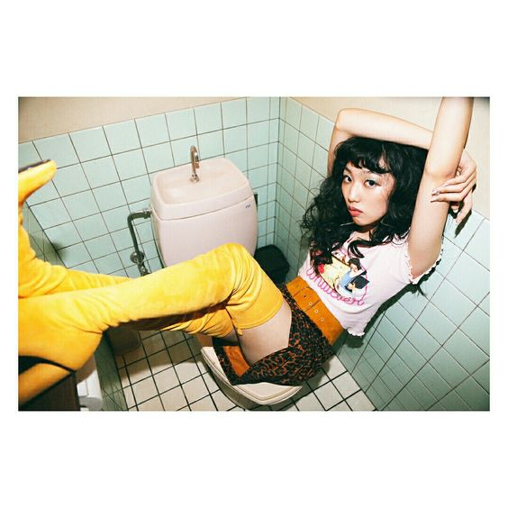 a woman is posing in a bathroom. she is wearing a pink and white tshirt, a leopard print skirt, yellow thighhigh boots, and a yellow belt. she is sitting on a toilet with her legs stretched out in front of her. she has dark hair and is looking at the camera. the bathroom has blue tiles on the walls and a white toilet and sink. the lighting is bright and artificial. the image is taken from a high angle.