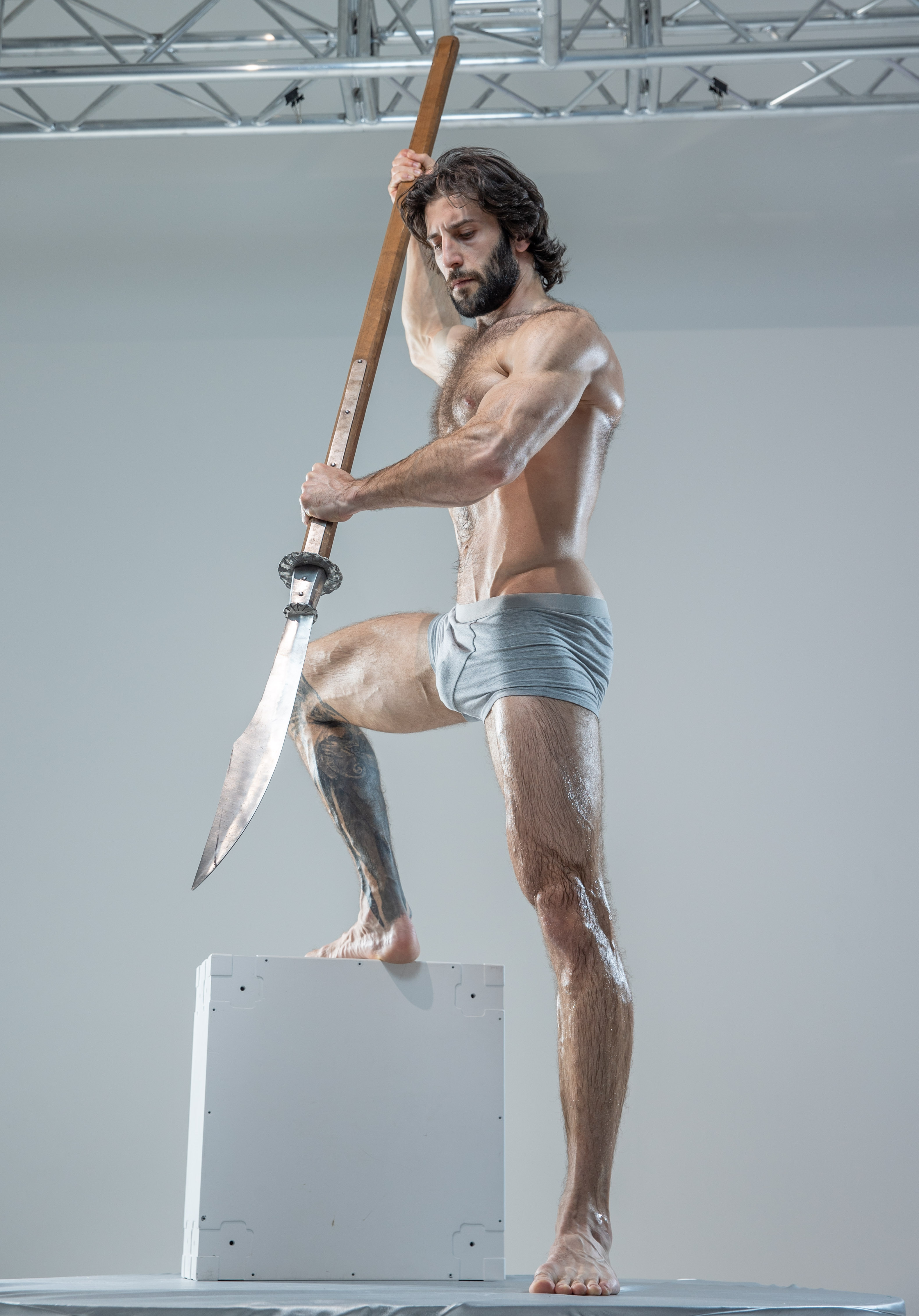 a muscular, bearded man with long, dark hair stands on a white cube in a studio setting. he is wearing only gray underwear and is covered in a light sheen of oil. he holds a large spear with a doublesided blade aloft in his right hand, his gaze focused downward. the background is a plain white wall, and the lighting is bright and artificial, highlighting the man's physique and the texture of his skin.