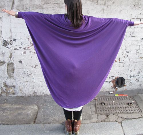 a woman standing on a street facing away from the camera, wearing an oversized purple shirt with her arms outstretched to the sides. the woman is wearing black tights and brown boots. a drain is visible on the street in the background.