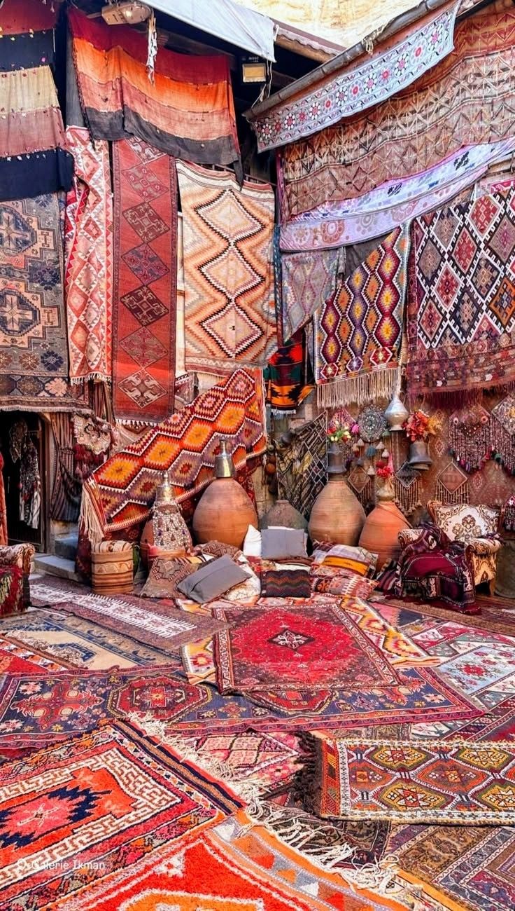 an image showing a shop or market stall selling a variety of colorful rugs, carpets, and textiles. the display is visually dense, with rugs hung on the walls and laid out on the floor, creating a layered effect of different patterns and colors. various pottery items, cushions, and pillows are also displayed among the rugs. the overall impression is of a traditional moroccan or oriental bazaar filled with handcrafted goods.
