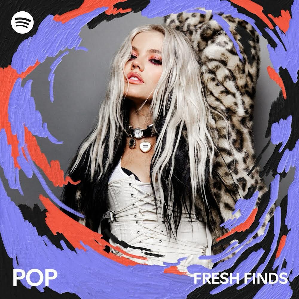 a woman with long blonde hair and black roots is featured on a spotify album cover. she wears a white corset, a black choker with a heart pendant, and an animal print coat. her makeup includes dark eye shadow and pink lipstick. the background is gray, and the album cover is decorated with paintlike streaks of lilac, orange, and black. the words "pop" and "fresh finds" are written in white at the bottom of the image. the spotify logo is in the top left corner.