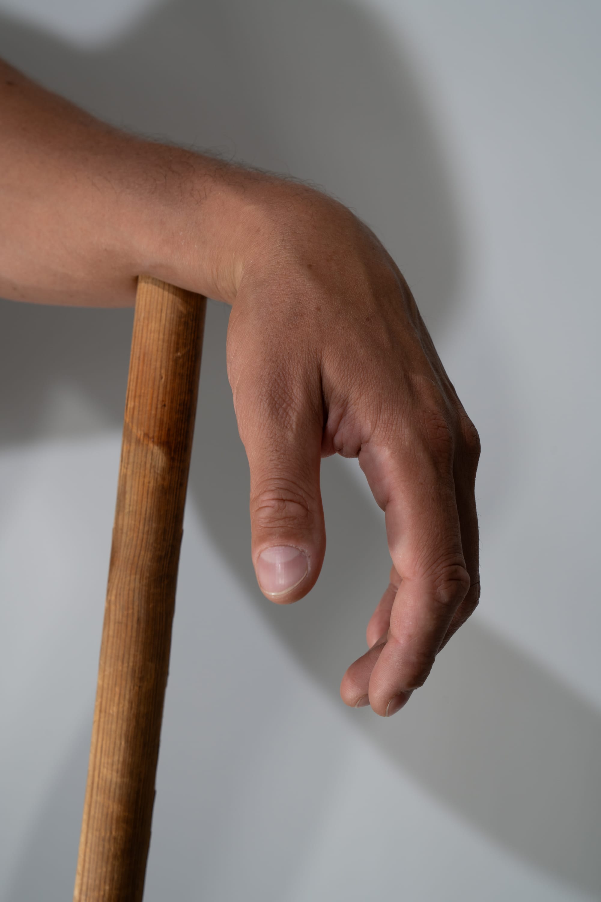 a closeup image of a man's hand gripping a wooden stick. the hand is illuminated by natural light, casting a shadow on the white background.