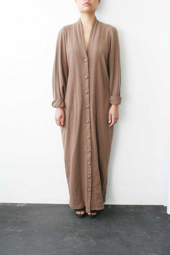a person with fair skin, wearing a light brown, longsleeved dress with buttons all the way down, is standing against a white wall. the dress is anklelength, and the person is wearing black sandals. they are looking down, and the floor is dark gray.