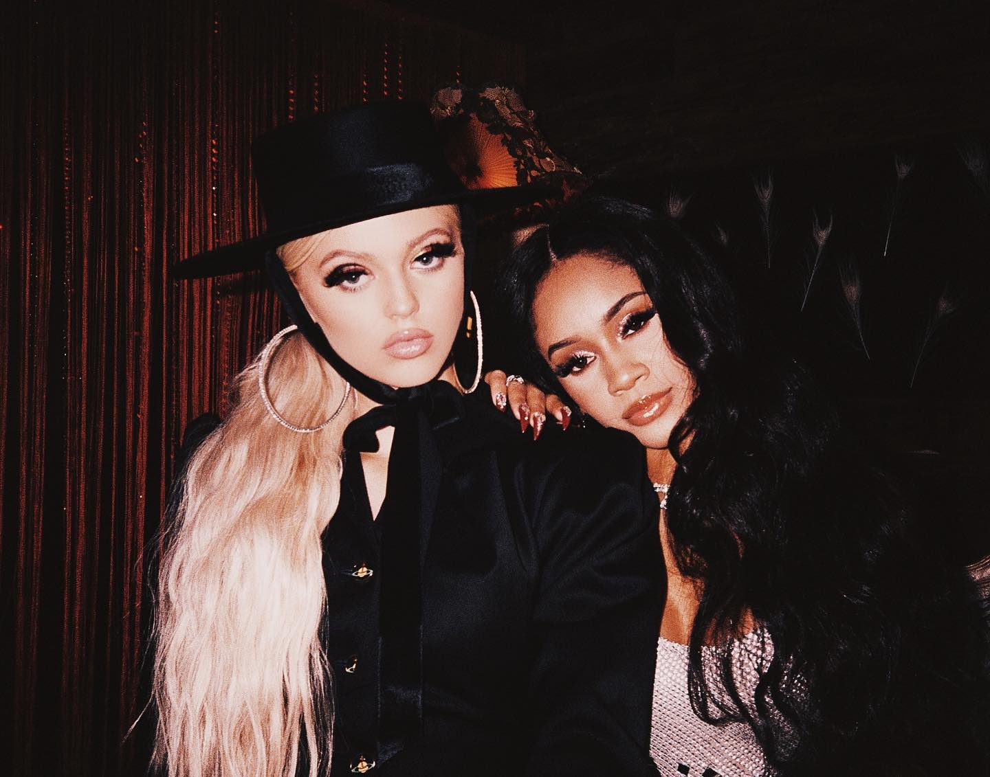 two women are posing closely together indoors. the woman on the left has long blonde hair, is wearing a black hat and suit, and has large hoop earrings. the woman on the right has long black hair, elaborate fingernail art, and is wearing a necklace. both women are wearing makeup, including long eyelashes and glossy lips.