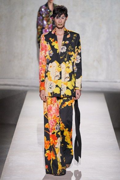 a full shot of a fashion model walking down a runway. the model is wearing a black pantsuit with a large floral print in shades of yellow, orange, and red. the pants are widelegged. the blazer is a classic style. the model has short, dark hair and minimal makeup. the background is a simple offwhite. the overall look is sophisticated and elegant.