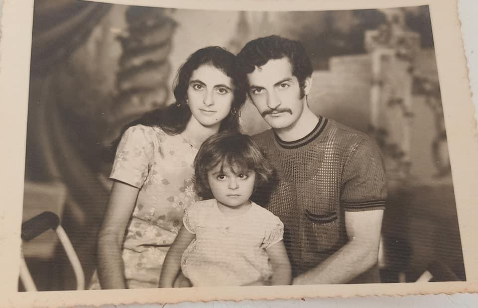 a sepiatoned vintage photograph shows a family portrait. a young woman with long dark hair and a floral dress sits next to a man with a mustache, wearing a textured shortsleeved shirt.  between them sits a small child with dark hair, also wearing a light colored dress. the family appears to be posed in a studio setting, with a blurred background suggesting some sort of backdrop. the photo has a slightly worn and aged look, typical of older photographs. the overall feel is nostalgic and reminiscent of family history.