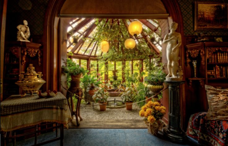 an ornate indoor space opens into a sunlit greenhouse. the room features antique furniture, including a table draped with a patterned cloth and a bookcase filled with books. statues are strategically placed, adding to the classic and elegant atmosphere. the doorway leads to a greenhouse filled with various plants and illuminated by spherical lanterns. the greenhouse has a glass roof and many windows, allowing natural light to flood the interior. there's a small fountain and a gravel ground in the greenhouse. yellow flowers in a pot are located near a black pedestal with a statue.