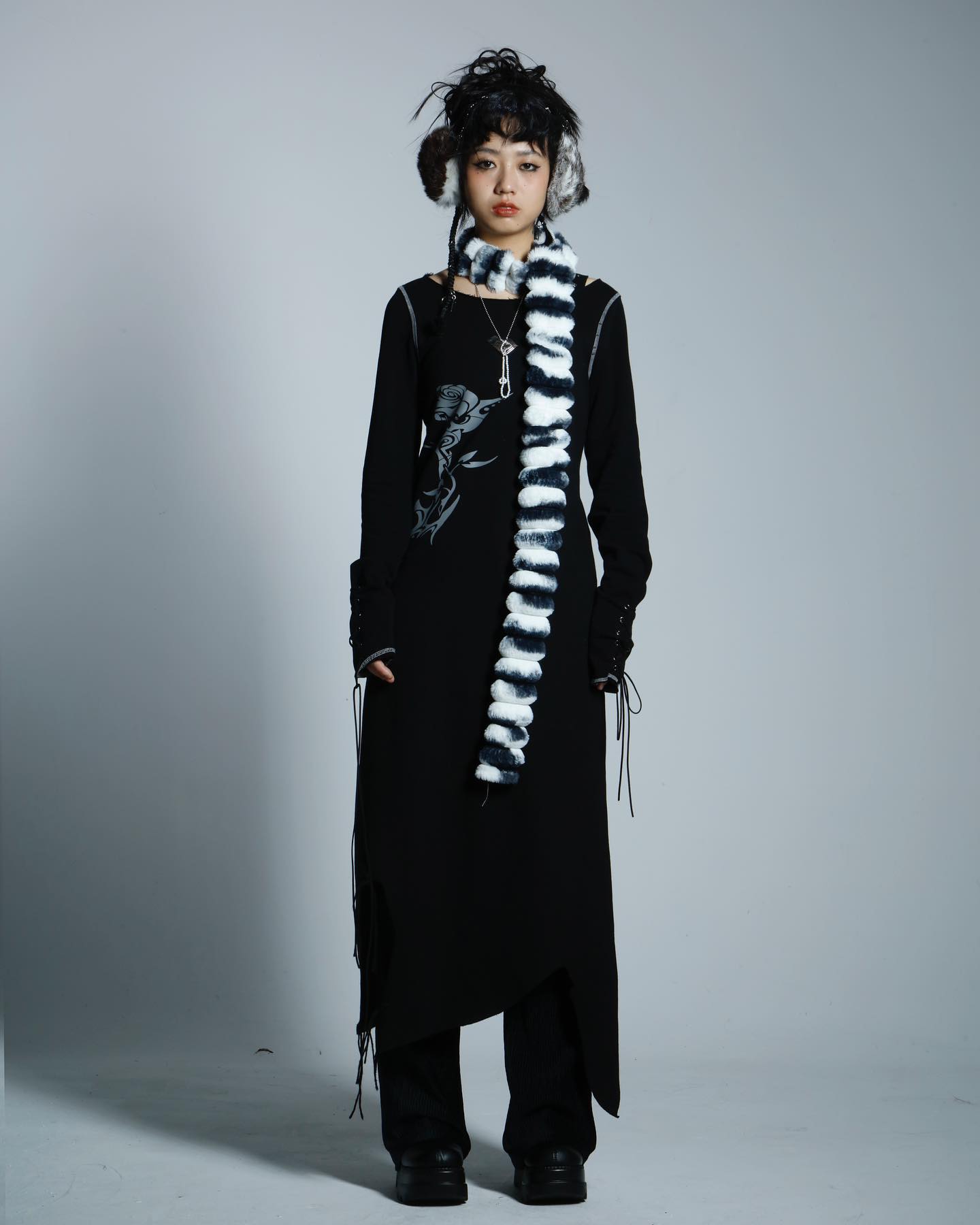 a fullbody studio shot of a young woman modeling a stylish outfit.  she's wearing a long black asymmetrical dress with side slits, paired with black pants and black boots.  she accessorizes with fur earmuffs in brown and white, and a long, vertically striped black and white fur scarf. her hair is dark and styled in a slightly messy fashion. the overall aesthetic leans towards gothic or alternative fashion styles. the background is a simple, light gray, which keeps the focus on the model and her outfit.