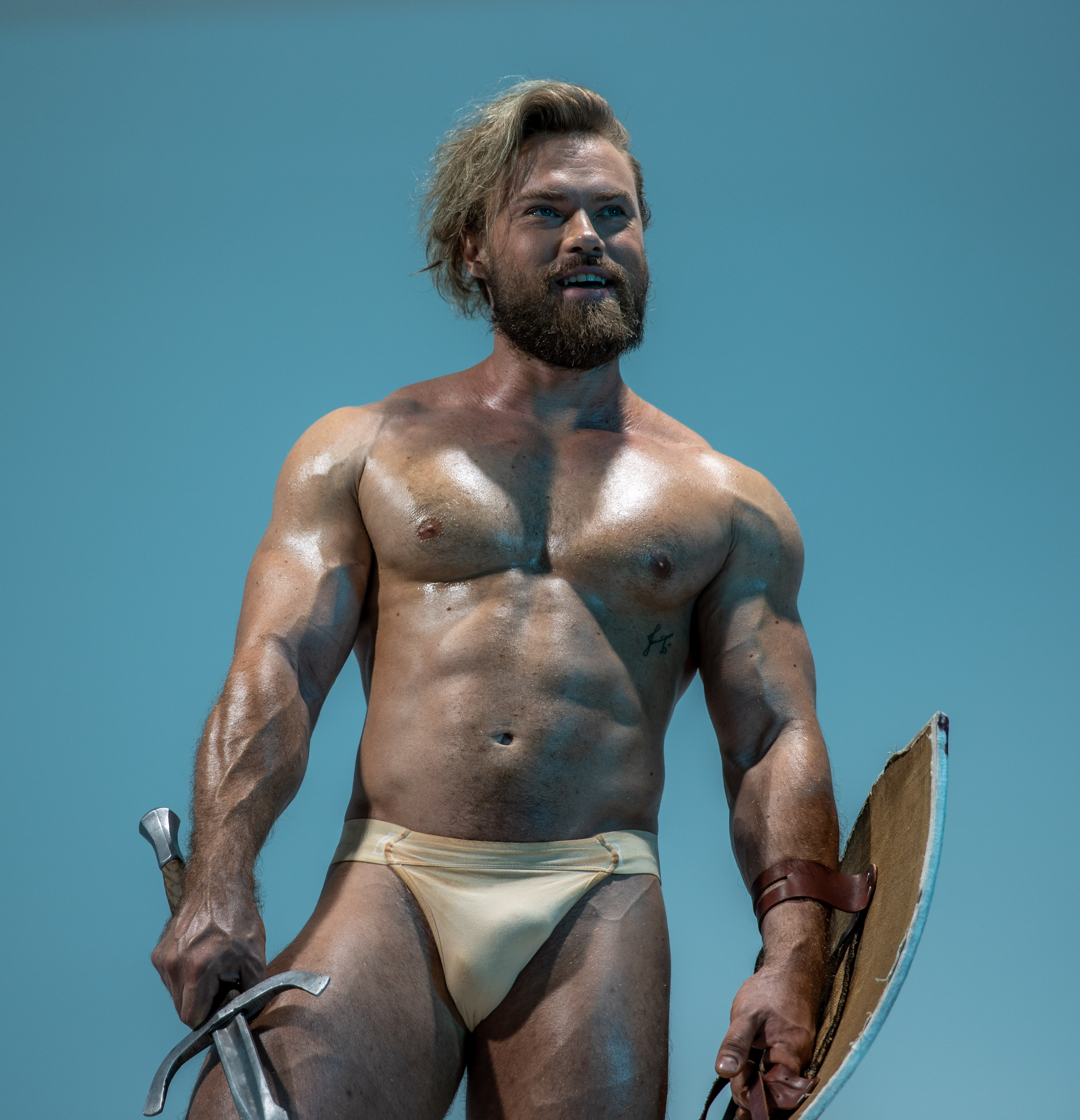 a muscular man with long blond hair and a beard poses in a studio setting against a light blue backdrop. he is wearing a loincloth and holding a sword in his right hand and a shield in his left, conveying the image of a warrior from ancient times. his physique is illuminated by studio lighting, enhancing his strong features.  the low angle perspective adds to his powerful stance.