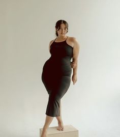 a young woman with long dark hair is posing in a black sleeveless midi dress. she is standing in a studio setting with a white background. her hair is down, and her makeup is minimal. she is looking at the camera with a confident expression.