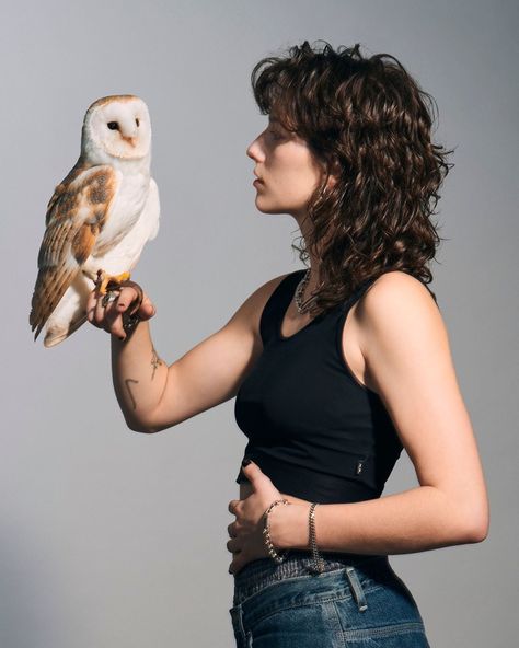 a young woman with shoulderlength curly hair stands in profile against a light gray background, gently holding a barn owl on her left hand. she's wearing a simple black tank top and blue jeans. her gaze is fixed intently on the owl, highlighting a sense of quiet connection between them.  the soft lighting emphasizes the textures of the owl's feathers and the woman's skin and hair. the image evokes a sense of peace and harmony with nature.