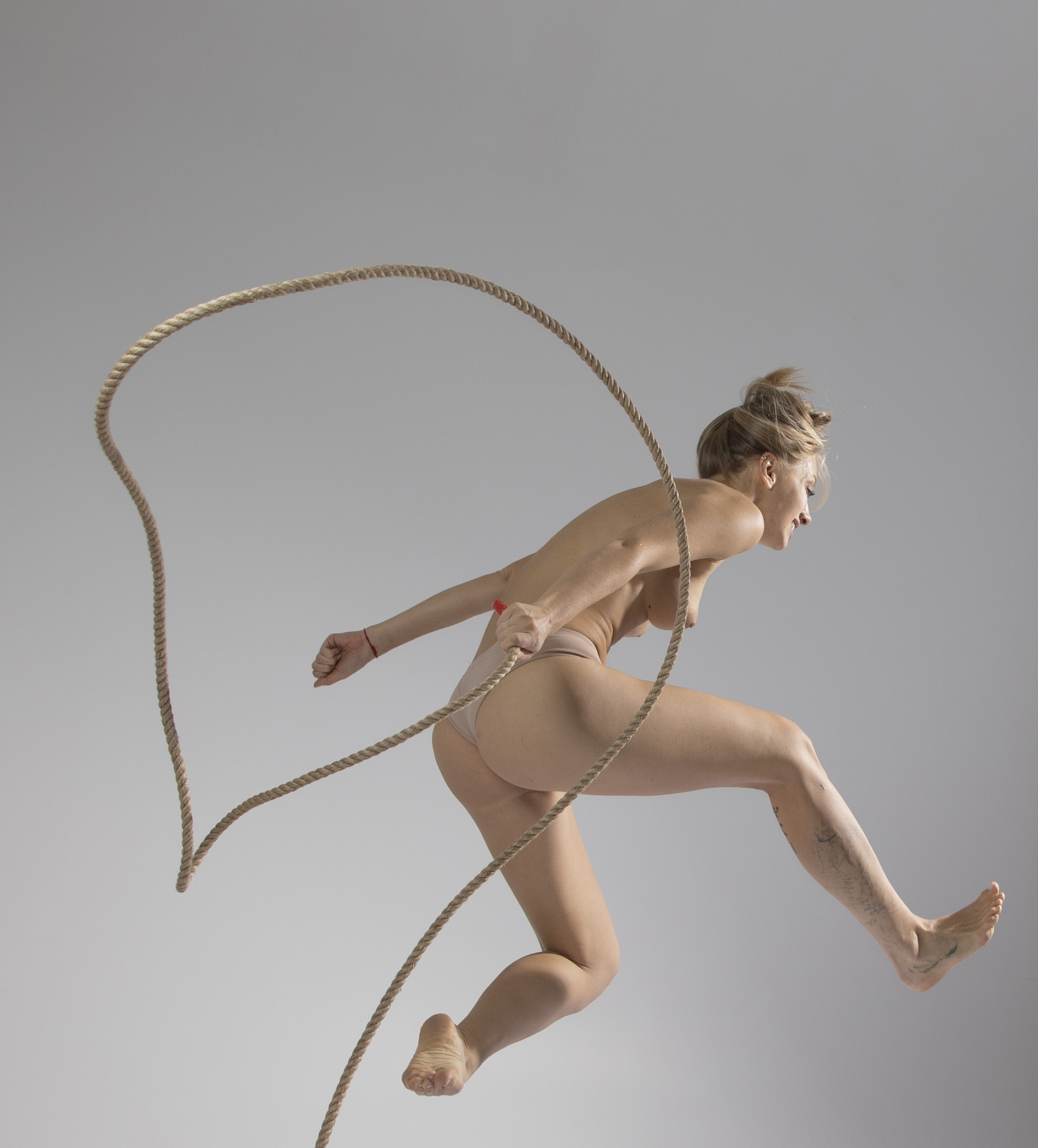 a nude woman is captured midair jumping rope in a studio setting. she is wearing what appears to be neutraltoned activewear and is barefoot. the rope forms a dynamic loop around her as she jumps, with a light gray backdrop providing a clean and minimalist background. the image emphasizes athleticism, movement, and a sense of physical activity.