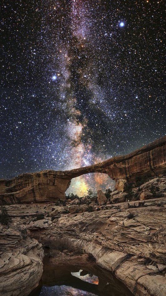 a stunning view of the milky way galaxy stretching across the night sky above a natural rock arch in a desert canyon. the arch is illuminated by the moonlight, creating a beautiful contrast against the dark sky. the stars are bright and clear, and the milky way is a breathtaking sight. the landscape is rugged and beautiful, with the rock formations adding to the majestic nature of the scene.
