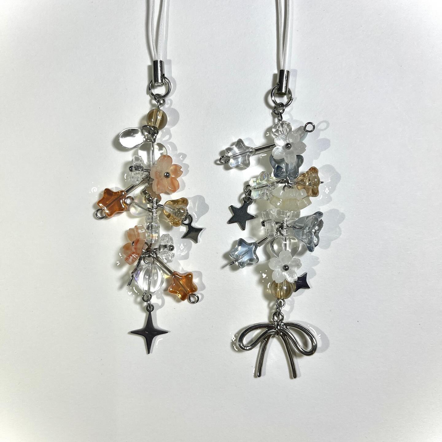 two handmade charm straps with bead and metal accents. the strap on the left features orange and peach flower beads, clear beads, and metal star pendants. the strap on the right features white and blue flower beads, clear beads, and a metal bow pendant. the straps are hanging from a white cord with a silver fastener.
