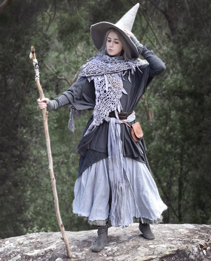 a woman dressed as a witch stands on a rock in a forest. she wears a tall grey witch hat, a long grey dress with layers and fringe, a knitted grey shawl, a brown belt with a small leather pouch, and grey boots. she holds a wooden staff in her left hand and her right hand rests on her hat. the background is a dense forest with varying shades of green.