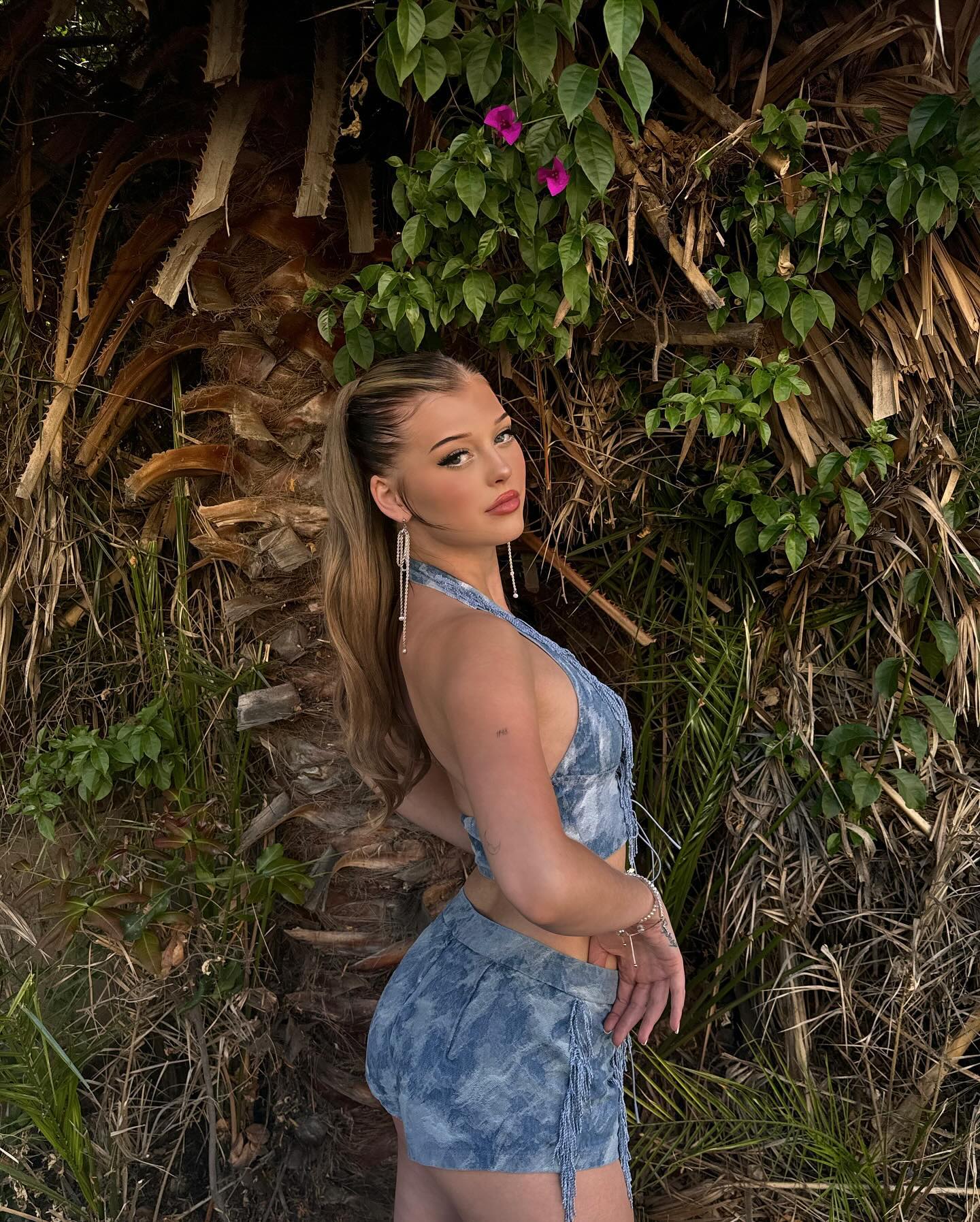 a woman with blonde hair stands in front of a tropical backdrop of leaves, flowers, and a tree. she is wearing a denim halter top and shorts with fringe. she is wearing jewelry, including earrings and bracelets. she is posing and looking over her shoulder.