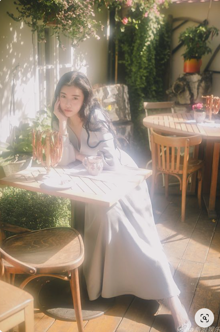 a woman in a light blue dress sits at a table in a sundrenched cafe. she rests her head on her hand and looks at the camera. the cafe has wooden tables and chairs and is decorated with plants. the lighting is soft and natural, creating a warm and inviting atmosphere.