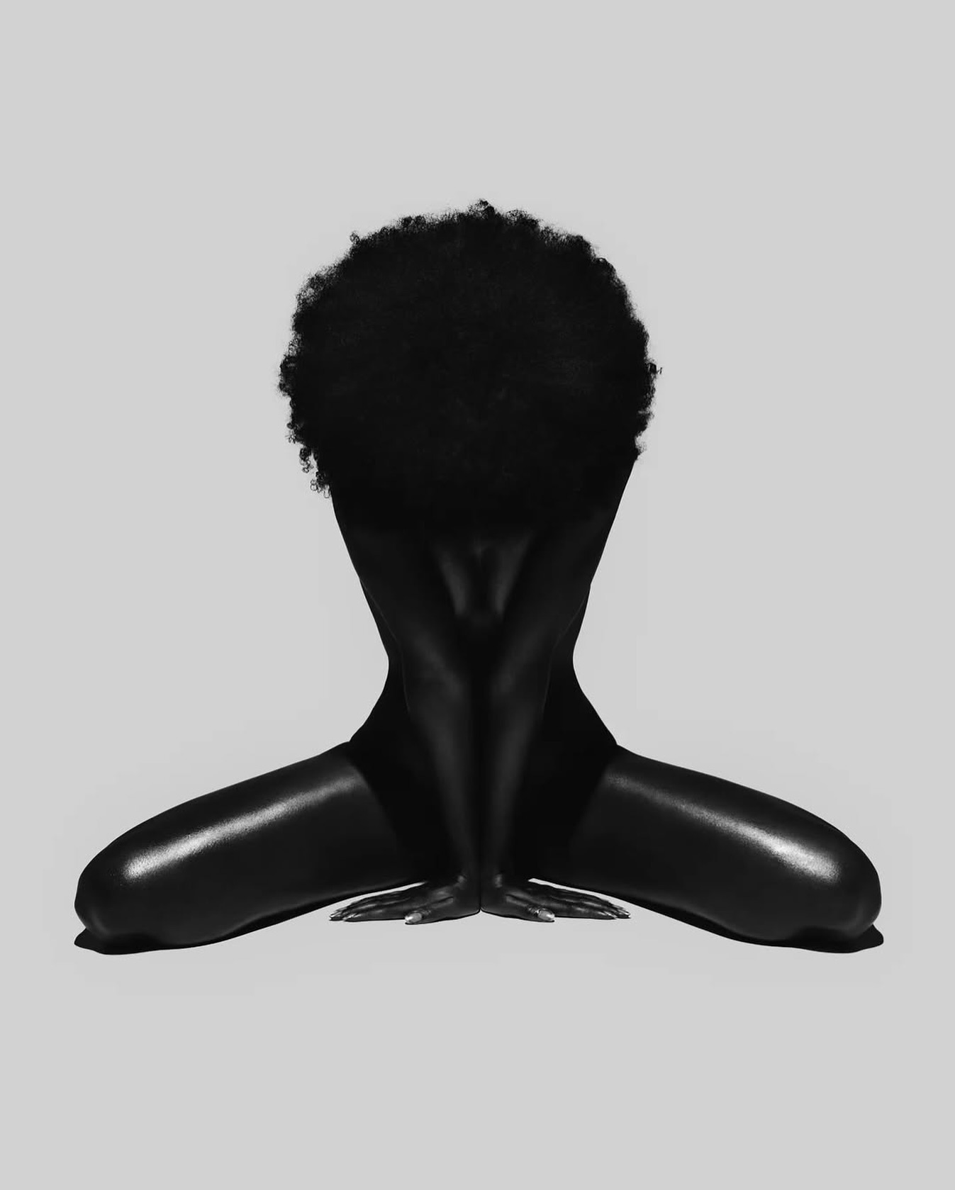 the image is a striking monochrome photograph of a darkskinned woman with a large, voluminous afro.  she is seated in a crouched position, her body presented in a frontal view. her arms are folded and her hands rest gently on the floor.  the lighting and photography technique creates strong contrasts and highlights the curves and shapes of her body, making it appear almost sculptural. the overall aesthetic is minimalist and artistic, emphasizing the beauty of the subject's form and her natural hair. the background is a plain, neutral gray, directing all attention to the woman.