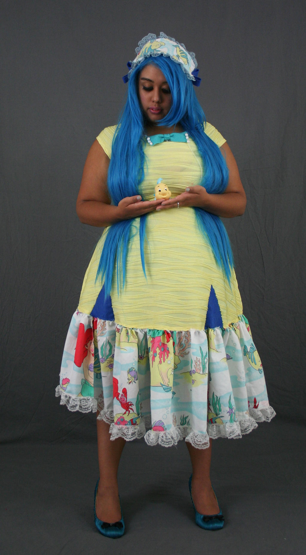 a full shot of a young woman cosplaying as a character, possibly inspired by disney's "the little mermaid". she has vibrant blue hair and is wearing a yellow dress with a seathemed print and white lace trim. the dress is cinched at the waist and has a ruffled skirt. the woman is holding a small yellow figurine (possibly flounder) in her hands. she's wearing a blue headband and teal heels, and she's posed against a gray backdrop. the overall style evokes a cute, whimsical, and slightly nostalgic aesthetic.