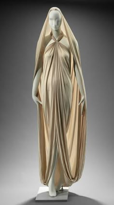 a white mannequin is wearing a long, draped dress. the dress is made of a sheer, flowing fabric and is cinched at the waist. the dress is very elegant and sophisticated. the mannequin is standing on a white platform. the background is a light gray.