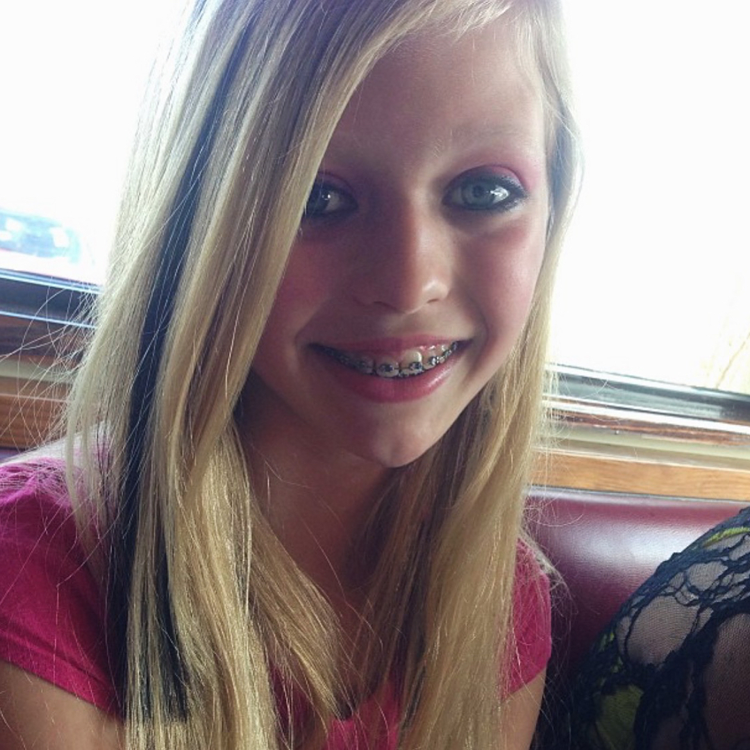 a young girl with blonde hair and braces is smiling in a closeup portrait. she is wearing a pink shirt and has black highlights in her hair. the background shows a window and a table. she also appears to be wearing makeup.