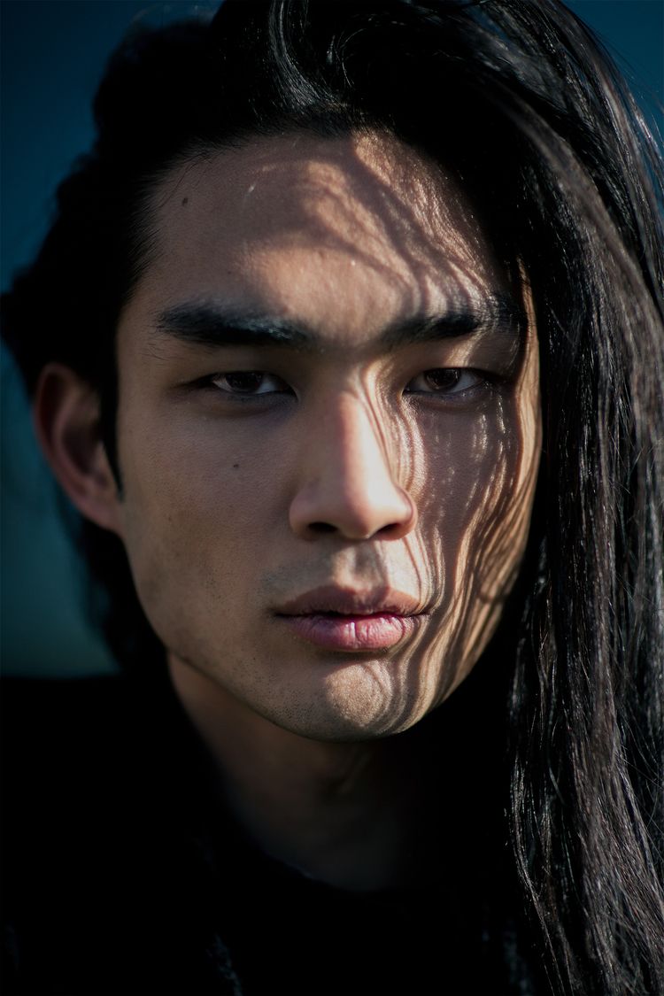 a closeup portrait of an asian man with long dark hair. shadows play across his face, creating a dramatic effect. he looks directly at the camera with a serious expression.