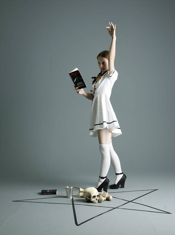 a young woman in a white dress and kneehigh socks stands within a pentagram drawn on a gray floor. she holds a book in her left hand and gestures with her right hand raised. her expression is serious and focused. outside the pentagram lay a black book, a metal chalice, and a human skull with a bone resting on top. the lighting is soft and artificial, illuminating the scene from the front.  the overall atmosphere is mysterious and suggestive of occult practices.