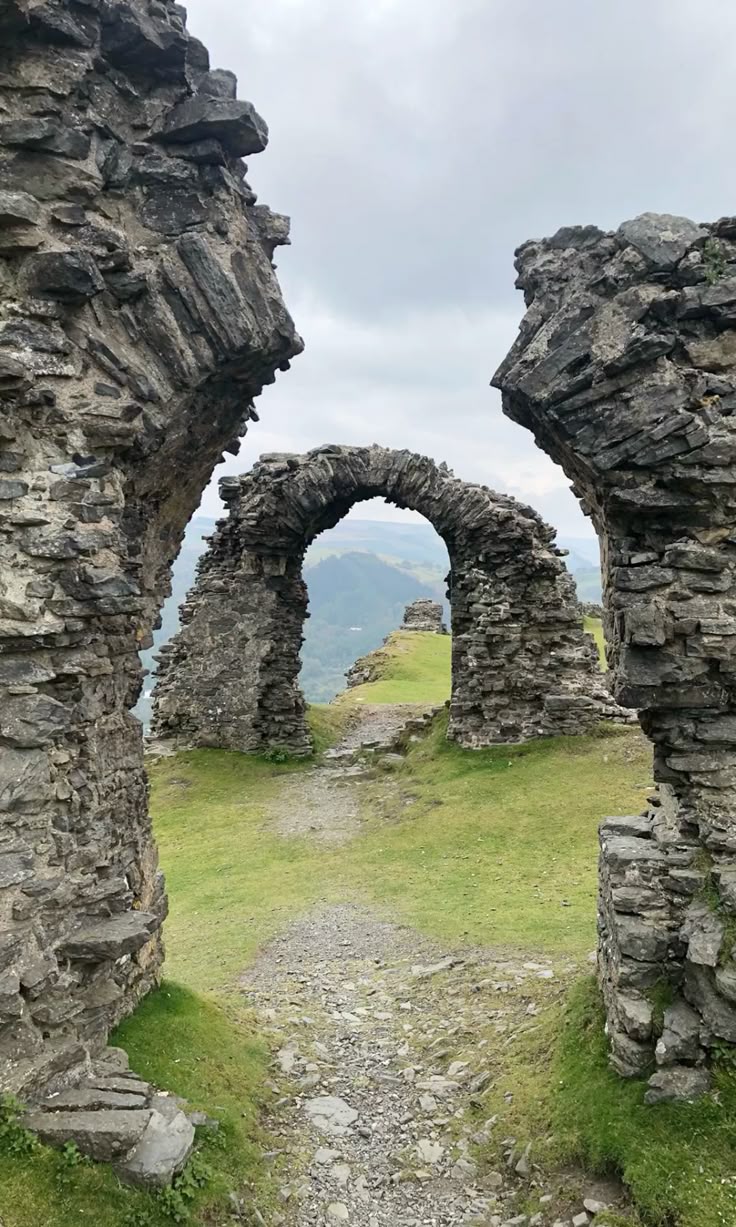 the image shows the ruins of a stone castle with three arches standing in a grassy area. a pathway leads through the arches, and in the background, there are mountains and a cloudy sky.