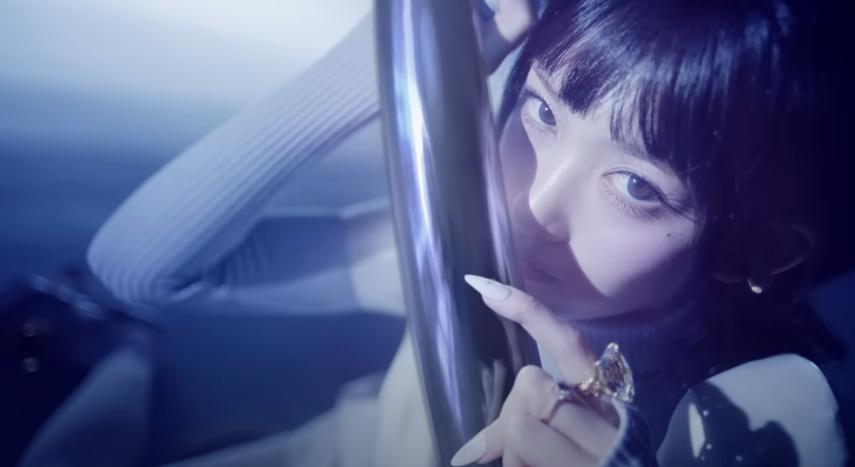 a closeup portrait of a young woman with dark hair and bangs. she's styled with dramatic eye makeup and long, decorated nails. she wears a silver ring on her index finger and a textured, longsleeved top. she holds onto a metallic pole, her gaze intense and directed towards the camera. the overall image is bathed in a cool, blue light, creating a moody and stylish atmosphere.