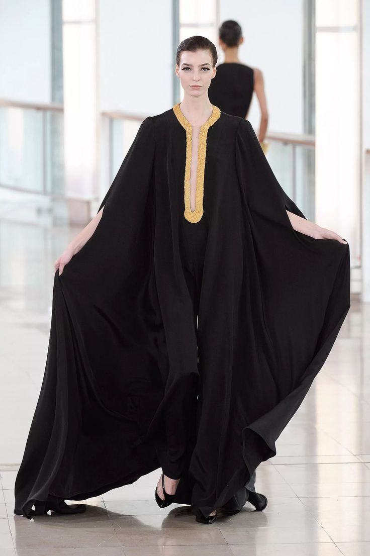 a female model walks down a runway in a black cape dress with gold accents. the model is posing for the camera, showing off the elegant and stylish design of the dress. the cape is flowing behind her, adding drama to the look. the model is walking in a professional manner, presenting the dress in the best light possible. the dress is likely part of a highfashion collection from a wellknown designer.  the model is  beautiful and stylish, demonstrating the beauty and creativity of the fashion world.  the background is white, giving the dress an even more dramatic feel.