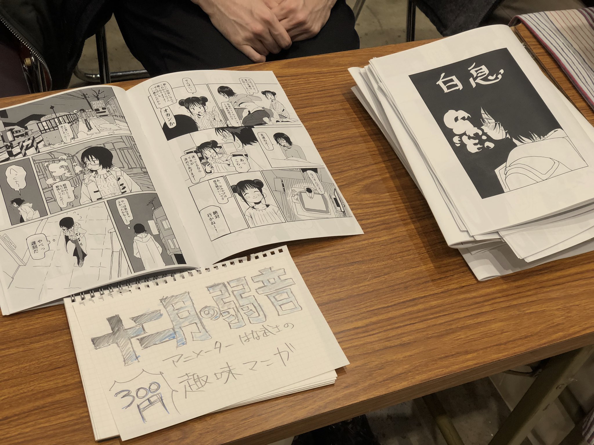 the image shows a wooden table with comic books on display. on the left, there's an open comic book with black and white illustrations and japanese text. in the middle, a handwritten sign advertises the comic for 300 yen. to the right, a stack of closed comic books with the title "白昼" are neatly piled up. two hands are subtly visible in the top left corner, implying the presence of a person behind the display. the setting appears to be an indoor event, likely a comic market where independent artists and publishers sell their work.