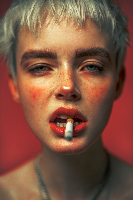 a closeup portrait of a young woman with short, bleached blonde hair. she is holding a cigarette between her lips, her expression is intense and somewhat sultry.  her skin is lightly freckled, and her makeup is subtly dramatic. the background is a deep, rich red, which contrasts with her pale skin. the overall mood of the image is edgy and artistic, with dramatic lighting creating a cinematic effect.