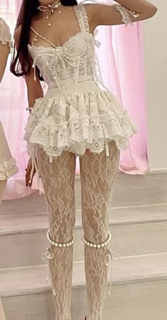 a woman in a white lace dress with a corset top, pearl necklace, and pearl bracelet is standing in front of a pink background. she is wearing white lace stockings. the woman is looking at the camera.